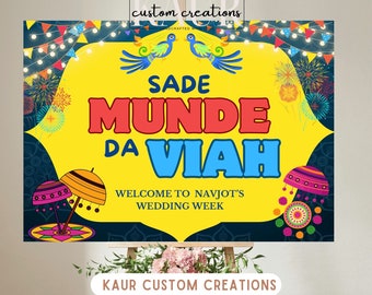 Op de afbeelding: Een geel en blauw bord met de tekst "Sade Munde Da Viah" en "Welkom bij Navjots huwelijksweek". Het bord is versierd met kleurrijke Indiase geïnspireerde ontwerpen, waaronder vogels, paraplu's en vuurwerk.