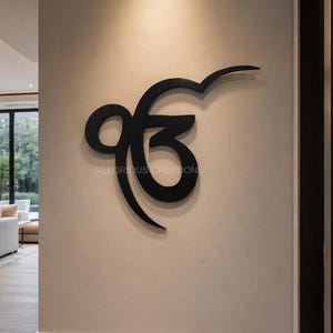 Puede incluir: Una pieza de arte mural negra con el símbolo Ik Onkar montada en una pared interior de color claro. El diseño presenta el carácter tradicional de la escritura punjabi con un acabado mate suave, creando un contraste con la pared.