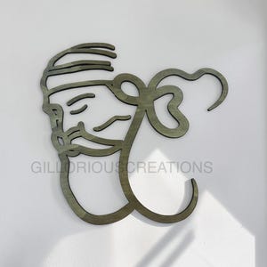 Könnte beinhalten: Ein Wandkunstwerk aus Holz, das ein stilisiertes Gesicht mit Bart und Turban in einem minimalistischen Design darstellt. Die Kunst ist in einem gedämpften Grünton gehalten und an einer weißen Wand befestigt. Der Text "GILLORIOUS CREATIONS" ist sichtbar.