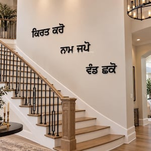 Op de afbeelding: Drie regels zwarte, driedimensionale muurkunst in Punjabi-schrift, gemonteerd op een lichte muur boven een houten trap. De tekst leest Kirat Karo, Naam Japo en Vand Chakko, met een strakke, vetgedrukte letterstijl die opvalt.