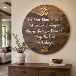 May include: A circular dark wood wall plaque featuring a gold Om symbol at the top, a gold lotus flower at the bottom, and white raised lettering displaying the Gayatri Mantra: Om Bhur Bhuvah Svah, Tat savitur Varenyam, Bhargo Devasya Dhimahi, Dhiyo Yo Nah Prachodayat.