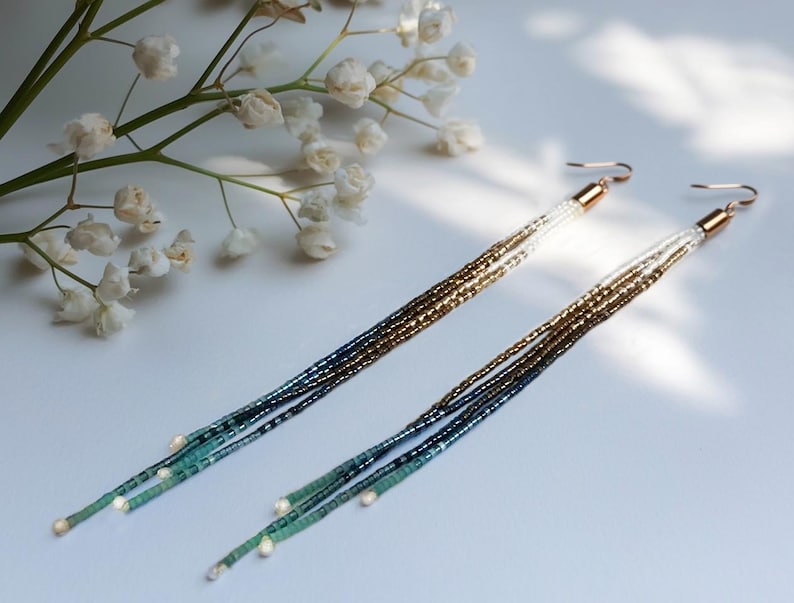 May include: A pair of long, beaded earrings with a gradient of colors. The earrings transition from gold to navy blue and teal, with small white beads at the ends. The ear wires are gold-colored. The earrings are displayed on a white surface.