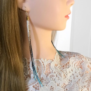 May include: Long, dangling earrings with a gradient of colors. The earrings feature a gold-colored top and transition from white to brown, then to dark blue and light blue beads. The earrings are worn by a person with long blonde hair.
