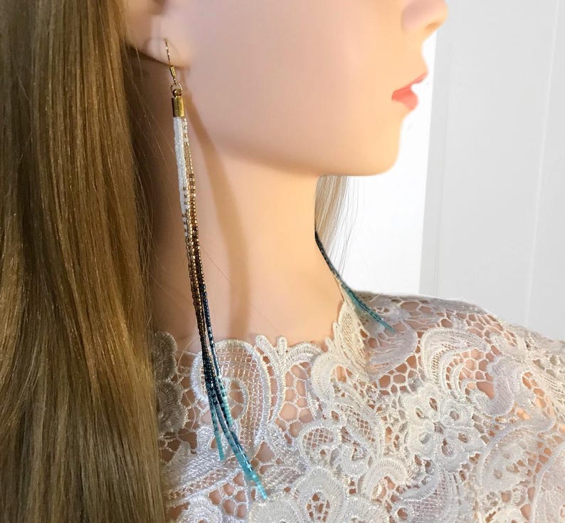 May include: Long, dangling earrings with a gradient of colors. The earrings feature a gold-colored top and transition from white to brown, then to dark blue and light blue beads. The earrings are worn by a person with long blonde hair.