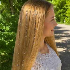 May include: Three thin, vertical hair strands featuring small, iridescent, round beads attached at intervals. The beads reflect light with blue and white tones, hanging straight down against long, straight, light-brown hair.