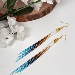 May include: A pair of handmade beaded earrings with a gradient of colors. The earrings feature gold-colored hooks and white, gold, brown, and blue beads. The earrings are long and dangle, with clear beads at the ends. The earrings are displayed on a white surface.