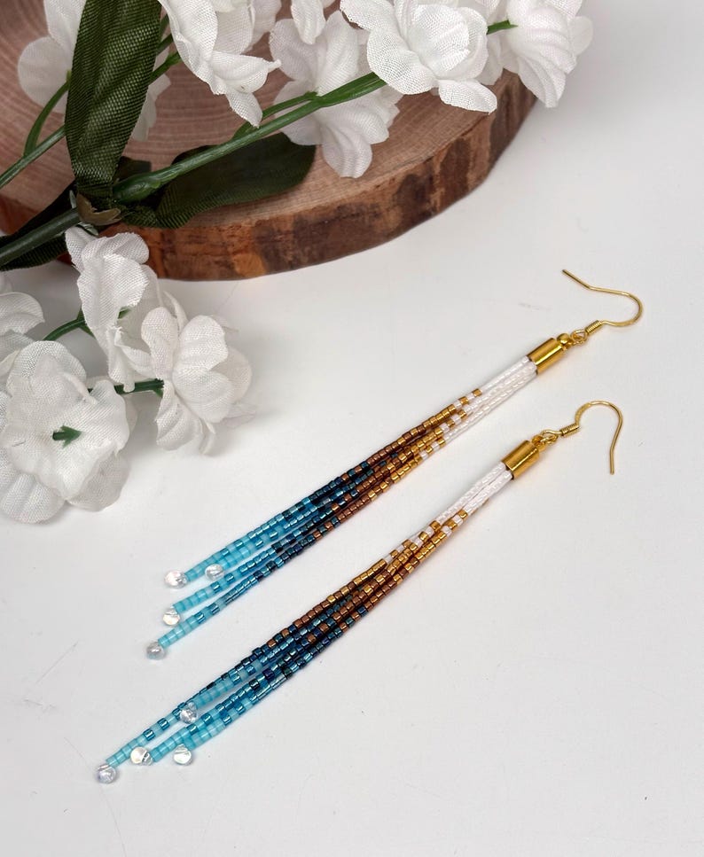 May include: A pair of handmade beaded earrings with a gradient of colors. The earrings feature gold-colored hooks and white, gold, brown, and blue beads. The earrings are long and dangle, with clear beads at the ends. The earrings are displayed on a white surface.
