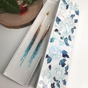 May include: Long beaded earrings in a white box with a floral design. The earrings have a gold, brown, and blue gradient. The box is white with a floral pattern in shades of blue and white.