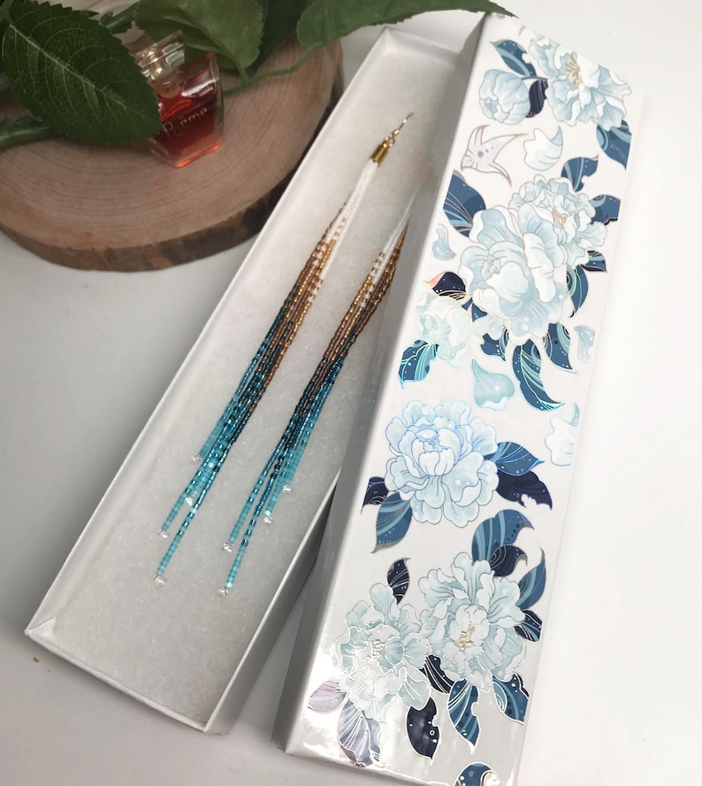 May include: Long beaded earrings in a white box with a floral design. The earrings have a gold, brown, and blue gradient. The box is white with a floral pattern in shades of blue and white.