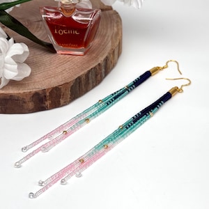 Könnte beinhalten: Ein Paar lange, perlenbesetzte Ohrringe mit Farbverlauf. Die Ohrringe wechseln von Marineblau zu Türkis, dann zu Rosa und enden mit klaren Perlen. Goldfarbene Akzente und Ohrhaken vervollständigen das Design. Die Ohrringe sind ca. 15 cm lang.