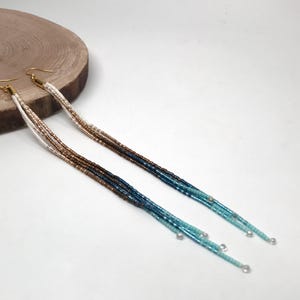 May include: A pair of long, beaded earrings with gold-colored hooks. The beads transition from white to brown, then to dark blue and turquoise. The earrings are displayed on a light background with a wooden disc.