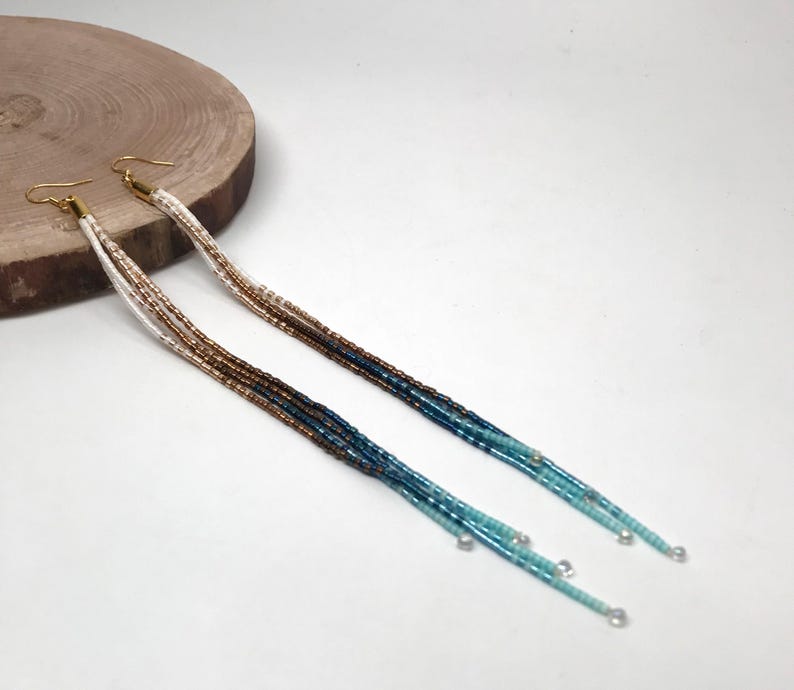 May include: A pair of long, beaded earrings with gold-colored hooks. The beads transition from white to brown, then to dark blue and turquoise. The earrings are displayed on a light background with a wooden disc.