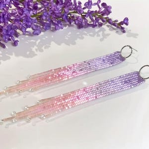 May include: Two beaded earrings in a pink to purple gradient, with a fringed design. They feature silver-tone circular ear wires. A sprig of purple flowers is visible in the background.