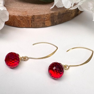 May include: A pair of gold-tone hook earrings featuring faceted, spherical red beads. The earrings have a curved, minimalist wire design that suspends the round, textured red stones, creating a bright and reflective appearance for the jewellery.