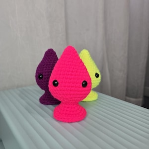 Puede incluir: Tres figuras hechas a mano con ganchillo en colores brillantes: morado, rosa intenso y amarillo. Cada figura tiene una base redondeada, un cuerpo en forma de lágrima y ojos de botón negros. Las figuras se exhiben sobre una superficie azul claro.
