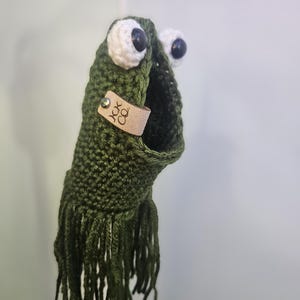 May include: Green crocheted frog with white eyes and black pupils. The frog has a brown leather tag with the text "JOK CO."