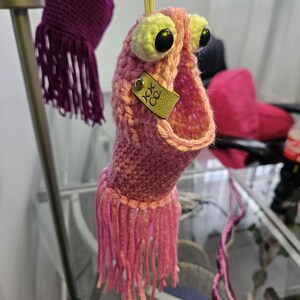 May include: A pink crocheted monster with large black eyes and yellow eyelids. The monster has a long, open mouth and a small gold tag that says "KK Co."