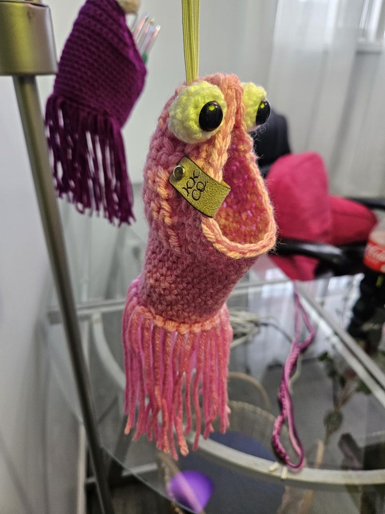 May include: A pink crocheted monster with large black eyes and yellow eyelids. The monster has a long, open mouth and a small gold tag that says "KK Co."
