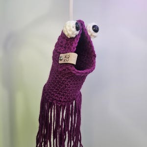 May include: Purple crocheted octopus with white eyes and black pupils. The octopus has a fringe of purple yarn and a small leather tag with the text "XOX CO."