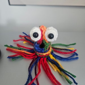May include: A vibrant, handmade crochet octopus toy with large googly eyes. The octopus is crafted in a rainbow of colours, including red, orange, yellow, green, blue, and purple. The tentacles are made of yarn and are various lengths.