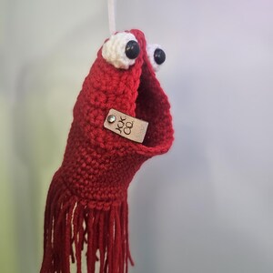 May include: Red crocheted hanging ornament with white eyes and black pupils. The ornament has a fringe at the bottom and a brown leather tag with the text "JOK CO."