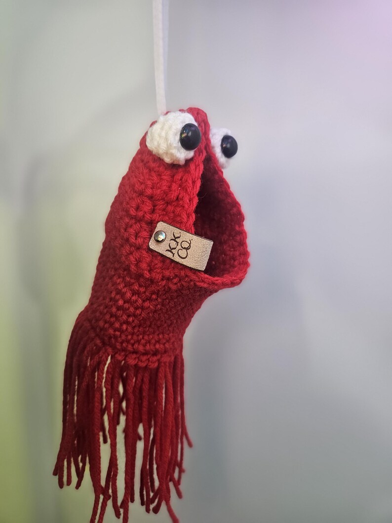 May include: Red crocheted hanging ornament with white eyes and black pupils. The ornament has a fringe at the bottom and a brown leather tag with the text "JOK CO."