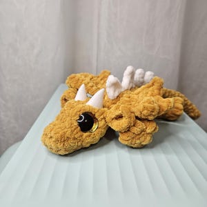 May include: A handmade, plush dragon toy in a golden-yellow color. The dragon features white horns and spine details, with large, black, glittery eyes. The toy is made of a soft, textured material.