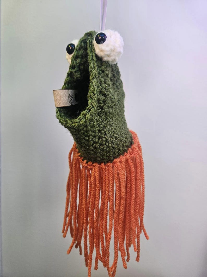 May include: Green crocheted frog ornament with large white eyes and a brown paper tag. The frog has a long orange fringe hanging from the bottom.