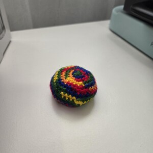 May include: A small, round, crocheted ball with a spiral design. The ball is a vibrant mix of colours, including red, yellow, green, and blue. This handmade item is likely a toy or decorative object.