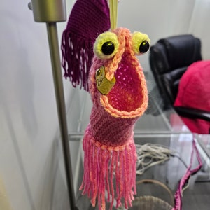 May include: A pink crocheted creature with large googly eyes and a fringe. The creature is hanging from a string and has a yellow tag with the word "Boo!" on it.