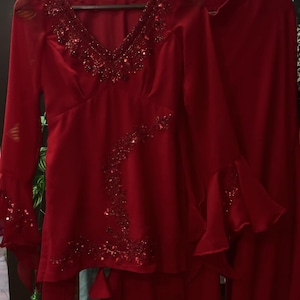 May include: A vibrant red two-piece outfit consisting of a long-sleeved tunic and matching trousers. The tunic features a deep V-neckline, flared sleeves, and intricate red sequin and bead embellishments along the neckline and side panel.