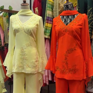 May include: Two matching outfits on mannequins, one pale yellow and one bright orange. Each set includes a long-sleeved tunic with flared sleeves, a V-neckline, and matching flared trousers. The tunics feature intricate beadwork and embroidery.