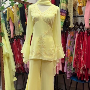 May include: A pale yellow two-piece outfit consisting of a long-sleeved tunic and matching wide-leg trousers. The tunic features a V-neckline, flared bell sleeves, and intricate tonal floral embroidery on the bodice. A matching scarf is draped around the neck.