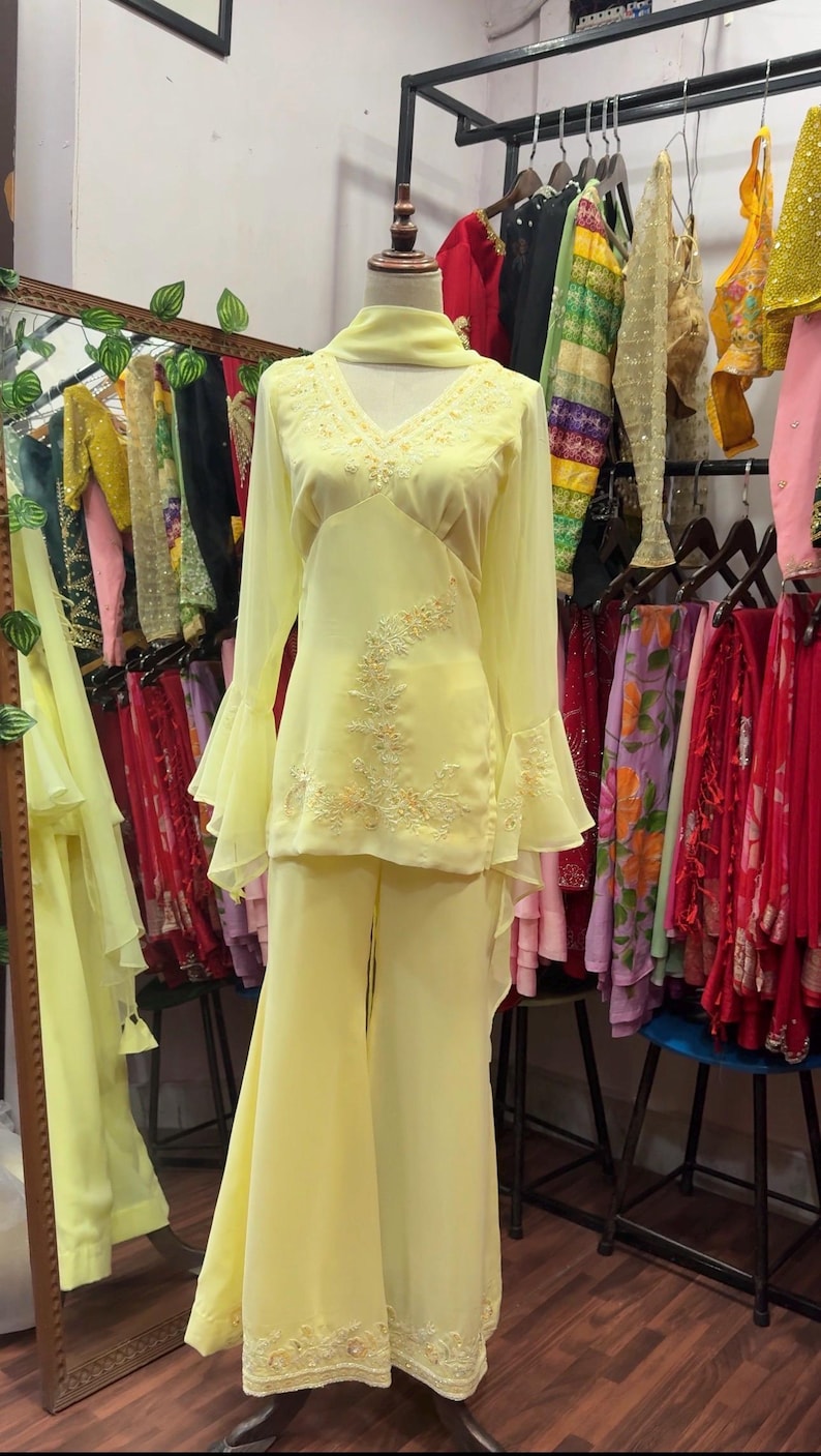 May include: A pale yellow two-piece outfit consisting of a long-sleeved tunic and matching wide-leg trousers. The tunic features a V-neckline, flared bell sleeves, and intricate tonal floral embroidery on the bodice. A matching scarf is draped around the neck.