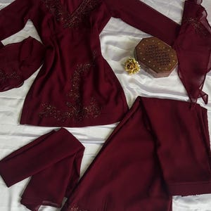 May include: A deep burgundy long-sleeved tunic and matching wide-leg trousers set. The tunic features a V-neckline with intricate dark beadwork and floral embroidery. The sleeves have flared cuffs with matching beadwork, and the trousers feature a wide embroidered hem.