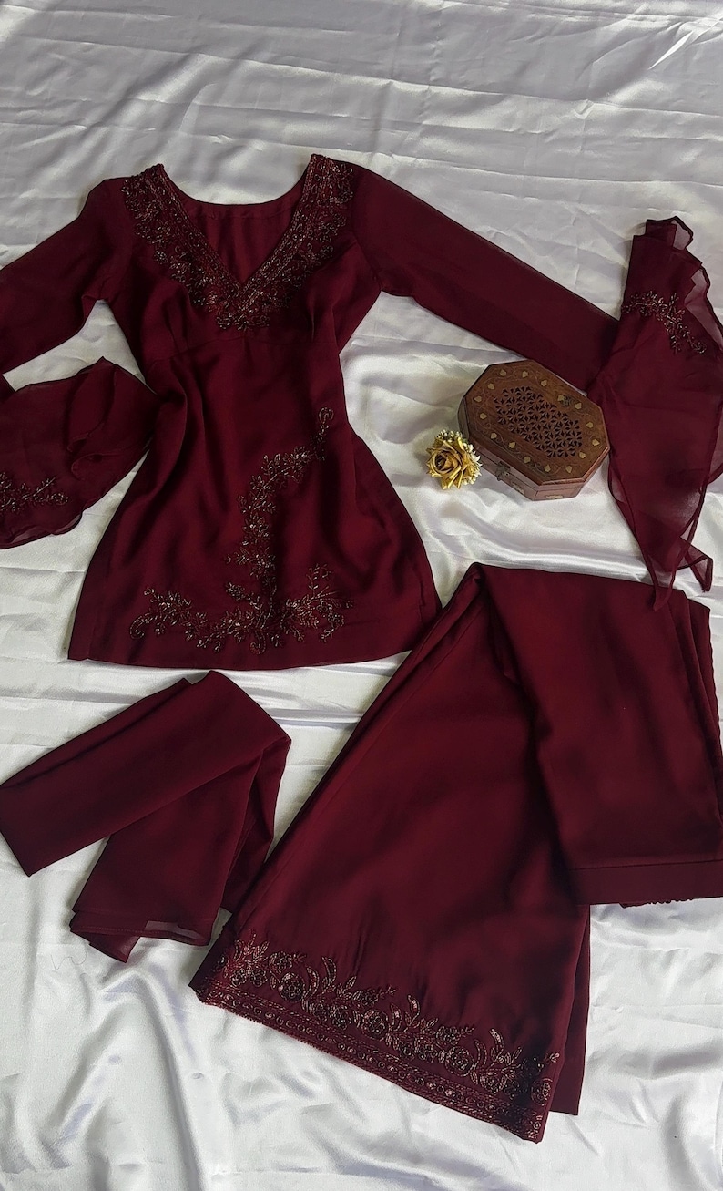 May include: A deep burgundy long-sleeved tunic and matching wide-leg trousers set. The tunic features a V-neckline with intricate dark beadwork and floral embroidery. The sleeves have flared cuffs with matching beadwork, and the trousers feature a wide embroidered hem.