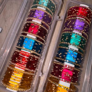 Op de afbeelding: Twee doorzichtige plastic containers gevuld met kleurrijke armbanden. De armbanden zijn in tinten goud, rood, blauw, paars en groen. Elke armband is versierd met kleine parels en reflecterende versieringen, wat een glinsterend effect creëert.