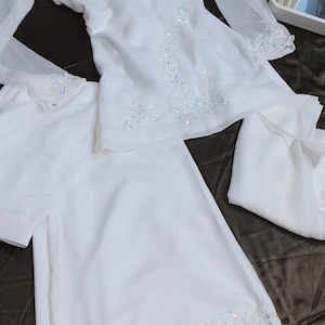 May include: A white long-sleeved tunic and matching trousers set featuring intricate white embroidery and iridescent sequins along the V-neckline, cuffs, and the hem of the trousers. The fabric is lightweight and semi-sheer with a flowing silhouette.