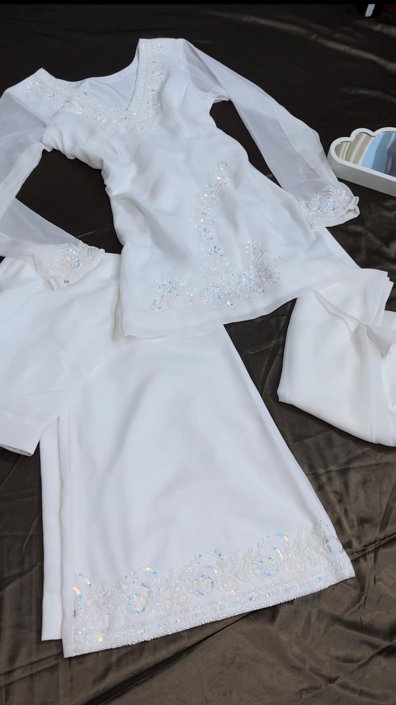 May include: A white long-sleeved tunic and matching trousers set featuring intricate white embroidery and iridescent sequins along the V-neckline, cuffs, and the hem of the trousers. The fabric is lightweight and semi-sheer with a flowing silhouette.