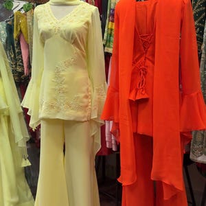 May include: Two outfits on mannequins: a pale yellow long-sleeved tunic with bell sleeves, matching flared trousers, and a scarf, alongside a bright orange long-sleeved tunic with a lace-up back, matching flared trousers, and a draped scarf.