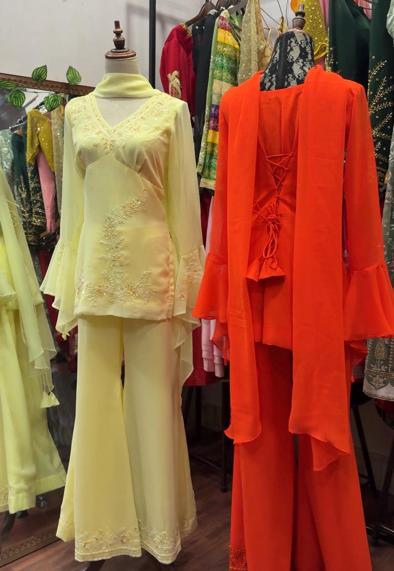 May include: Two outfits on mannequins: a pale yellow long-sleeved tunic with bell sleeves, matching flared trousers, and a scarf, alongside a bright orange long-sleeved tunic with a lace-up back, matching flared trousers, and a draped scarf.