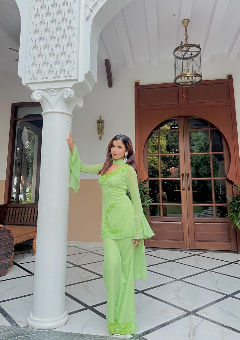 May include: A lime green two-piece outfit consisting of a long-sleeved tunic with flared cuffs and matching wide-leg trousers. The tunic features a high neckline, a cutout detail at the chest, and intricate tonal embroidery along the bodice and hem.