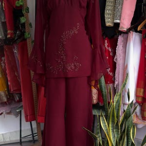 May include: A deep burgundy two-piece outfit consisting of a long-sleeved tunic and wide-leg trousers. The tunic features a V-neckline with intricate beadwork, bell sleeves, and a floral beaded design on the side. The trousers have beaded hems.