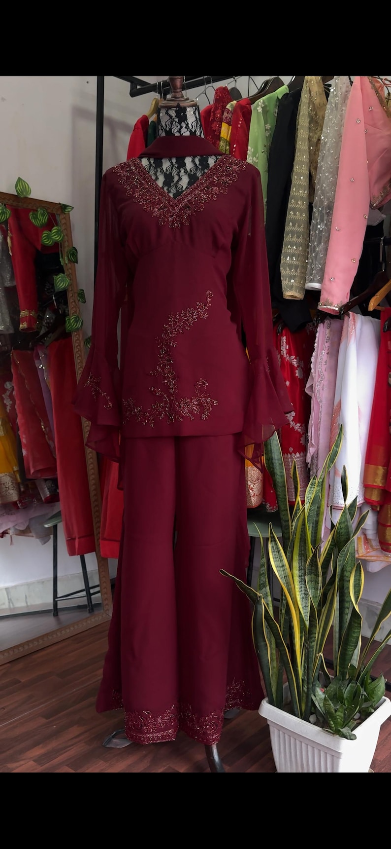 May include: A deep burgundy two-piece outfit consisting of a long-sleeved tunic and wide-leg trousers. The tunic features a V-neckline with intricate beadwork, bell sleeves, and a floral beaded design on the side. The trousers have beaded hems.