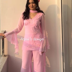 May include: A light pink two-piece outfit featuring a long-sleeved tunic with flared sleeves, a V-neckline, and intricate embroidery, paired with matching wide-leg trousers. The text Anjali's design is visible on the tunic front.