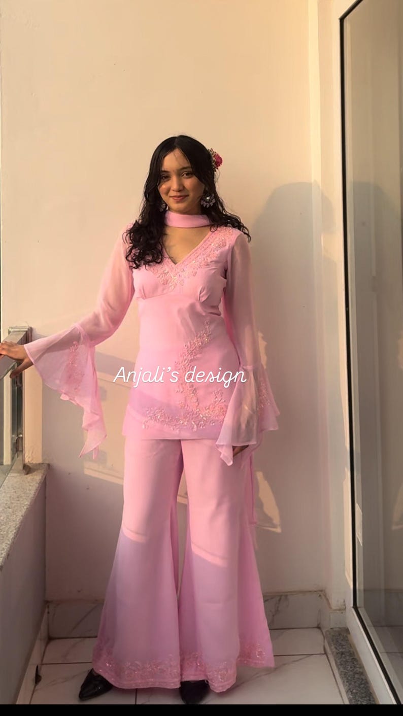 May include: A light pink two-piece outfit featuring a long-sleeved tunic with flared sleeves, a V-neckline, and intricate embroidery, paired with matching wide-leg trousers. The text Anjali's design is visible on the tunic front.