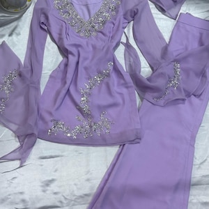 May include: A lavender long-sleeved tunic and matching wide-leg trousers set featuring intricate silver sequin and bead embroidery along the V-neckline, cuffs, and hem. The ensemble includes a coordinating lavender chiffon scarf or dupatta.