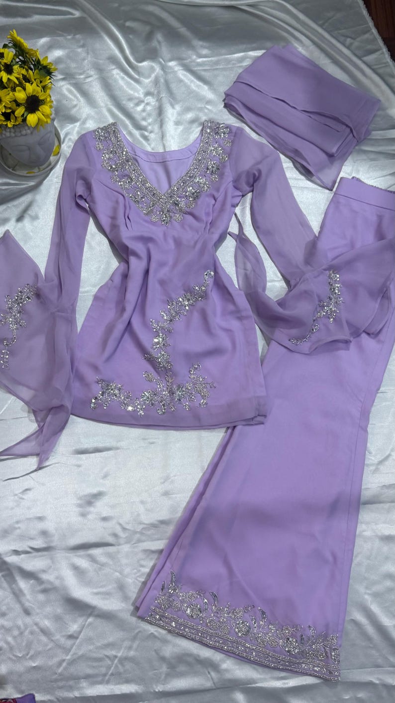 May include: A lavender long-sleeved tunic and matching wide-leg trousers set featuring intricate silver sequin and bead embroidery along the V-neckline, cuffs, and hem. The ensemble includes a coordinating lavender chiffon scarf or dupatta.