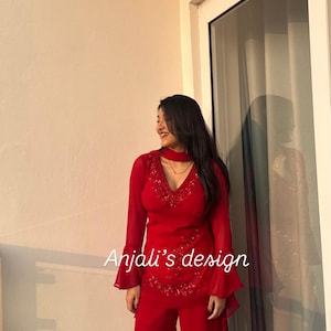 May include: A vibrant red two-piece outfit featuring a long-sleeved tunic with flared cuffs, a V-neckline with intricate red beadwork, and matching wide-leg trousers. The text Anjali's design is printed in white across the center of the tunic.