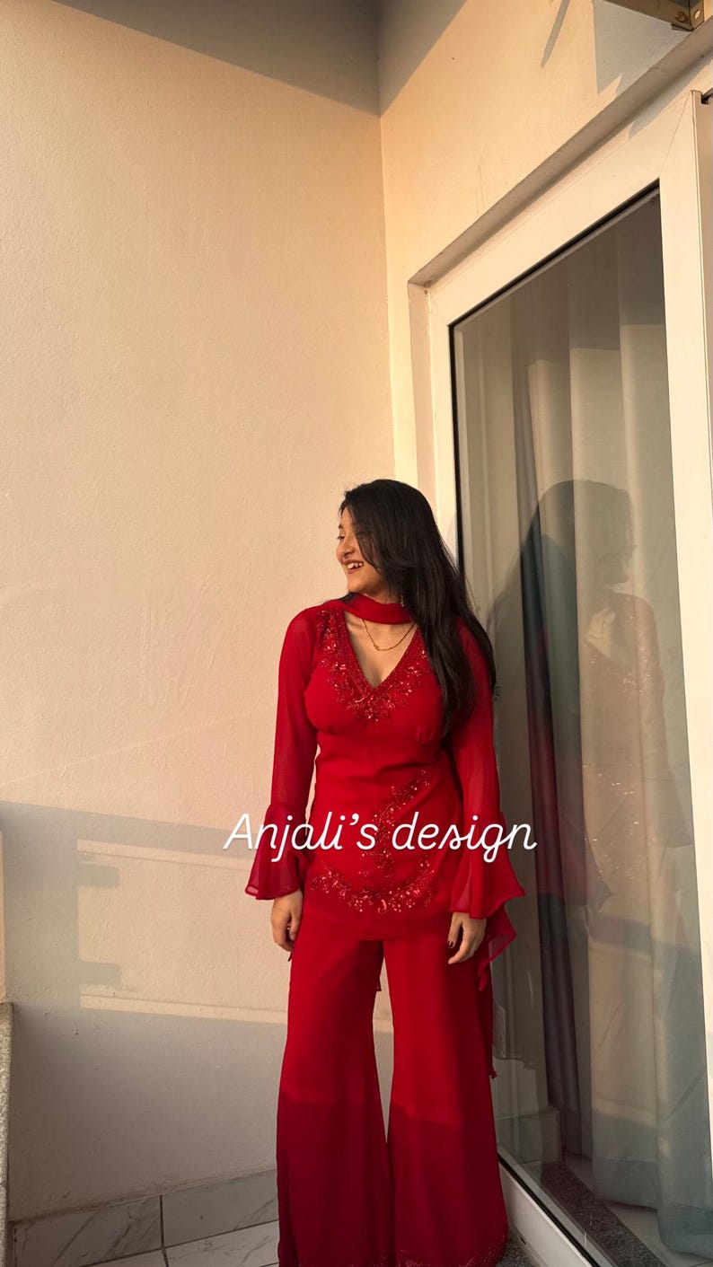 May include: A vibrant red two-piece outfit featuring a long-sleeved tunic with flared cuffs, a V-neckline with intricate red beadwork, and matching wide-leg trousers. The text Anjali's design is printed in white across the center of the tunic.